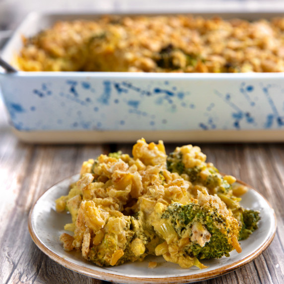 A plateful of broccoli-cheese casserole on a wooden table with the full casserole dish behind it.