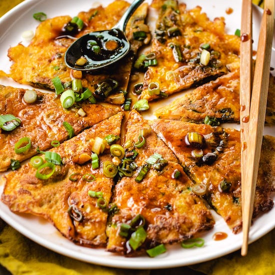Kimchi pancake with scallions on a white plate with chopsticks and a spoon with sauce