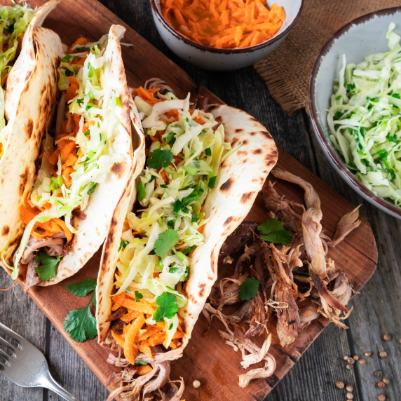 Pulled Lamb Tacos with Pickled Shallots, Cabbage Slaw, and Carrot Salad on wooden board.
