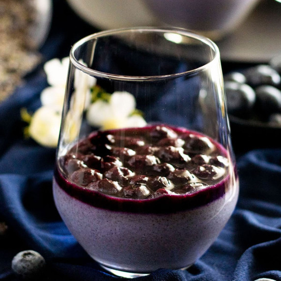 A glass of lavender and blieberry panna cotta with blueberry sauce on navy fabric.