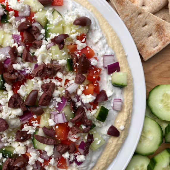 Layered hummus dip topped with feta, cucumbers, red onion, red pepper and olives on a white plate. 