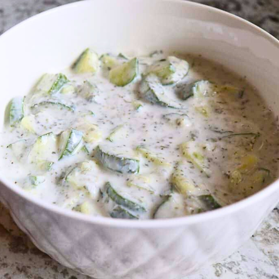 White ceramic bowl of chopped cucumbers in creamy, herb-flecked yogurt and garlic infused sauce