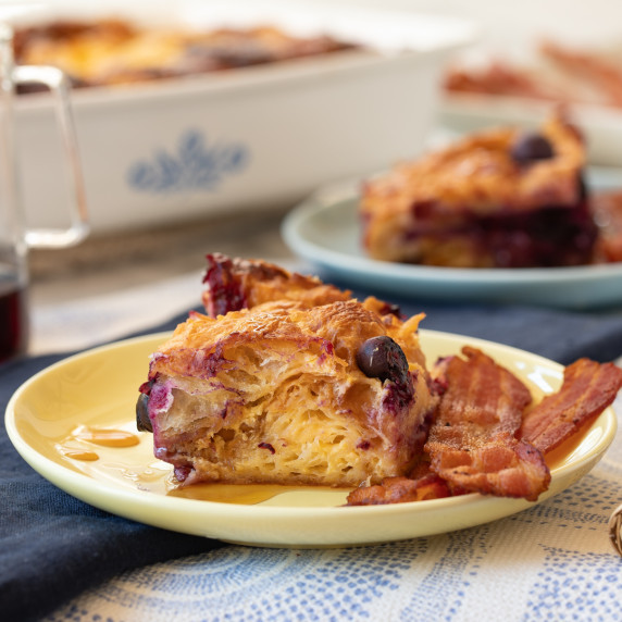 A slice of Lemon Blueberry Croissant Bake on a yellow plate with a side of bacon.