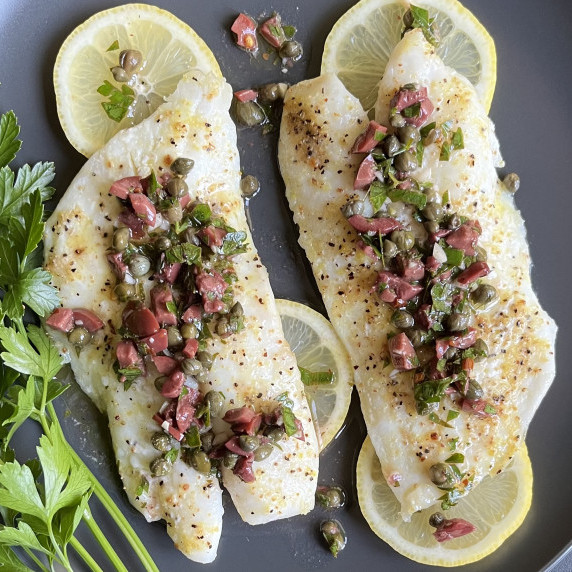 Flounder fillets topped with olives and capers on a black plate.