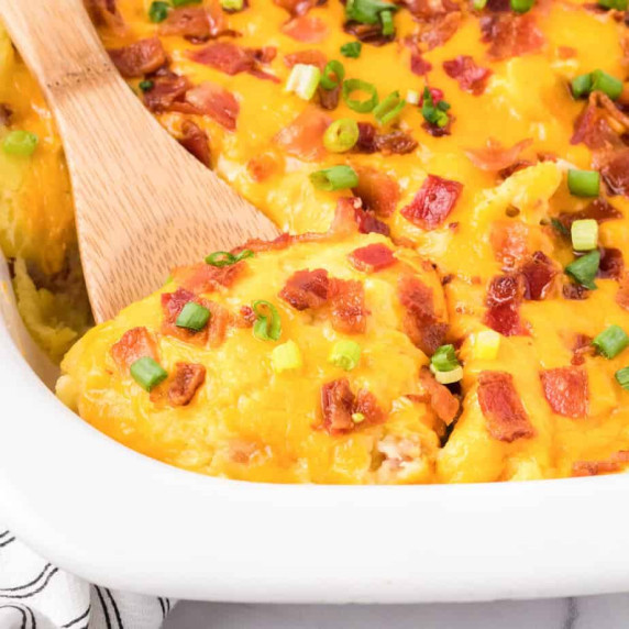 Square close up view of a spoon scooping mashed potato casserole covered in bacon and cheese.