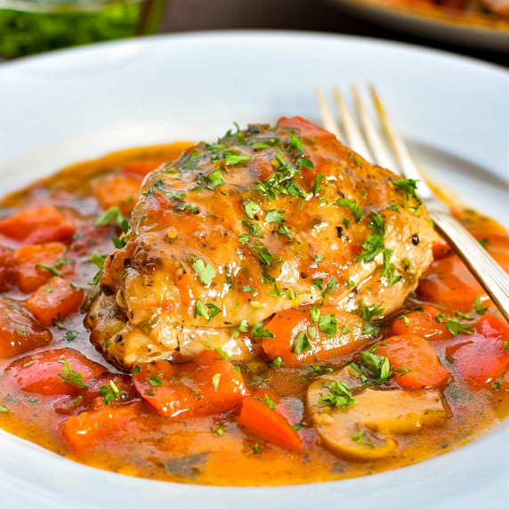 chicken cacciatore on a brown rimmed platter next to a large spoon and bowl of chopped parsley