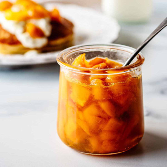 Glass jar of mango compote with a spoon on a marble surface