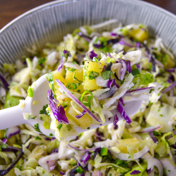Close up shot of Mayo Free Pineapple Slaw on white spoon with bowl of the slaw in the background