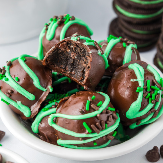 Chocolate Oreo truffles in a white bowl with green sprinkles with the top piece missing a bite.