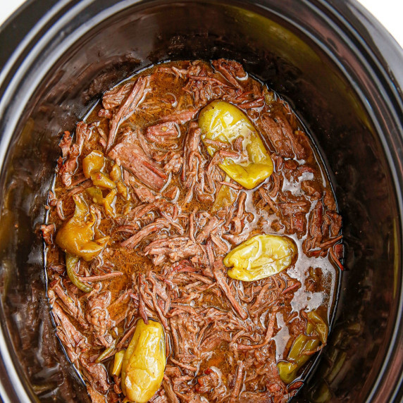 A slow cooker filled with shredded Mississippi pot roast beef, cooking liquid and peppers from above