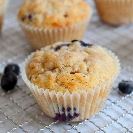 Close-up of a moist, golden blueberry muffin in a paper liner on a wire rack with fresh blueberries.