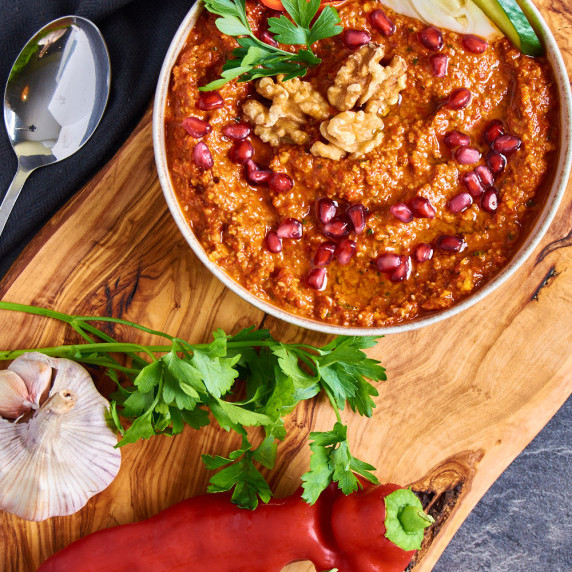 A bowl filled with creamy red Muhammara, topped with walnuts and pomegranate seeds