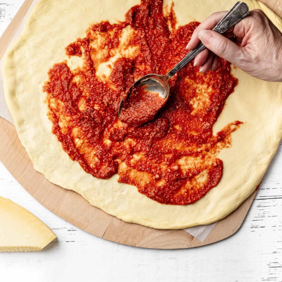 Pizza sauce being spooned onto shaped raw pizza dough