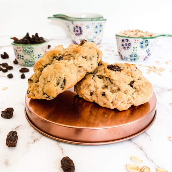 Two oatmeal raisin cookies on a copper platform with raisins and oats in measuring cups behind