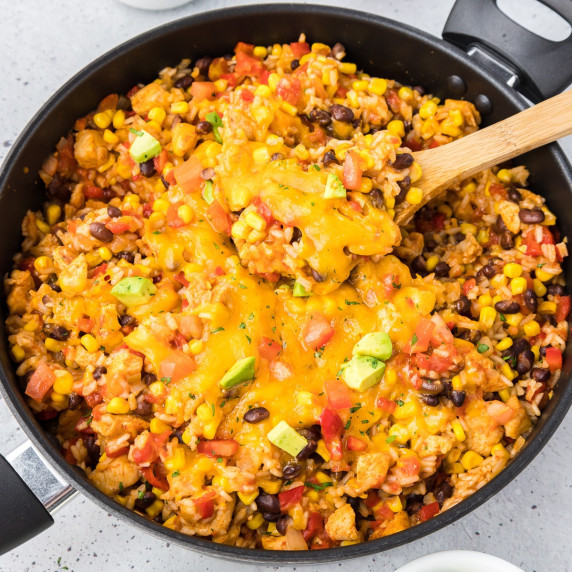 Cheesy Mexican rice, chicken and vegetables in a skillet with a wooden spoon scooping out a portion.