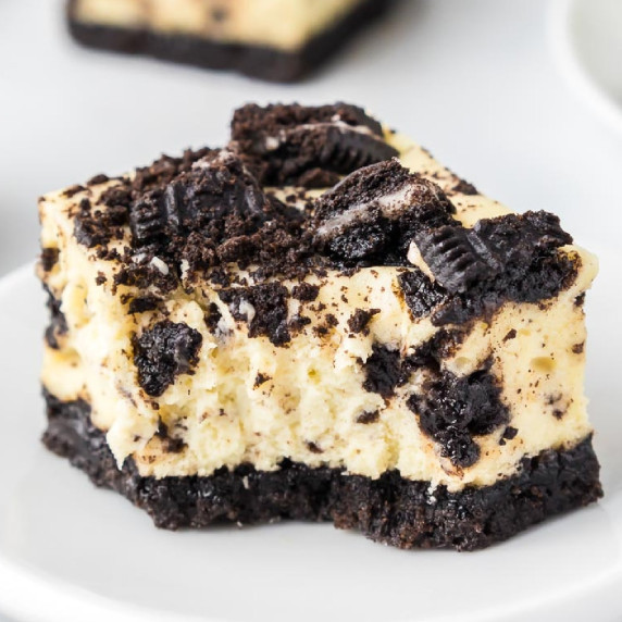 Oreo cheesecake bar on a plate topped with Oreo cookie pieces with a bite missing.