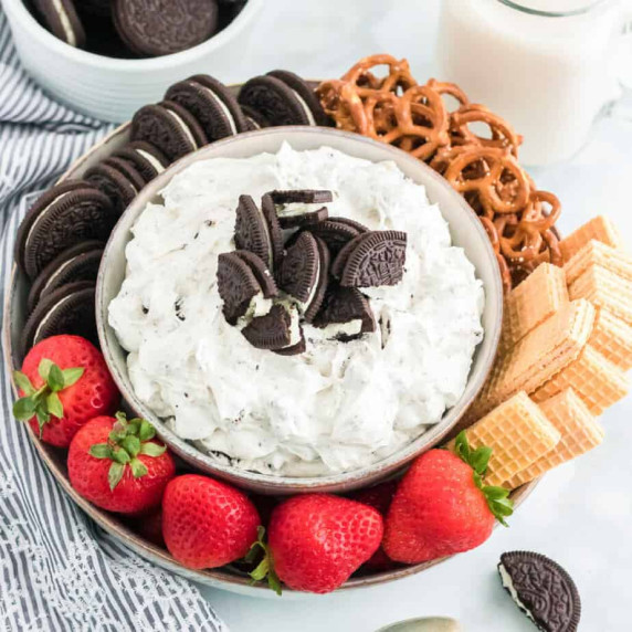 Close up of a bowl of dessert dip topped with Oreo cookies on a round platter with food to dip.