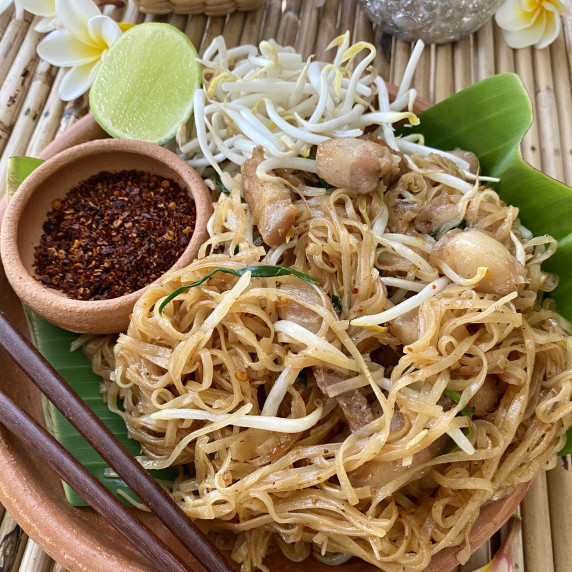 Pad mee korat (spicy pad Thai) on a banana leaf in a clay dish with wooden chopsticks.