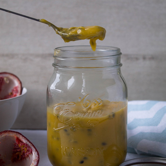passion fruit curd in a jar with a spoonful being lifted out surrounded by empty passion fruits