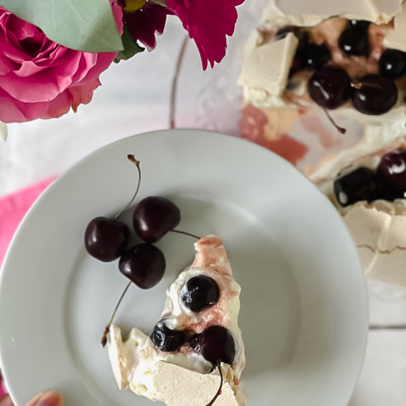 Slice of Pavlova topped with cherry on a white plate.
