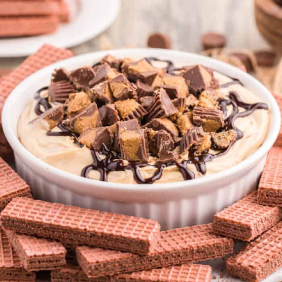  Cheesecake dip close up topped with peanut butter cups and chocolate in a bowl next to cookies. 