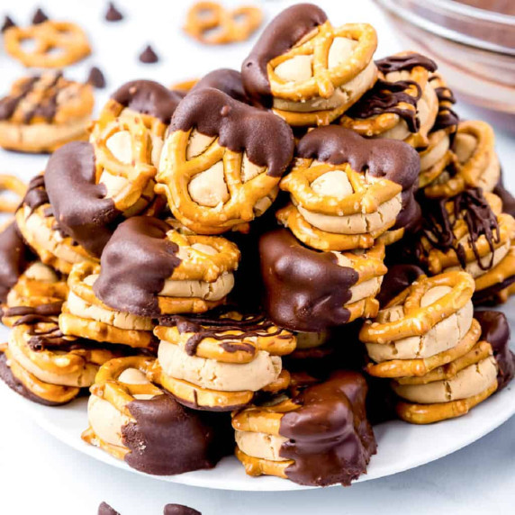 Peanut butter stuck between small pretzels dipped in chocolate stacked high on a plate from the side