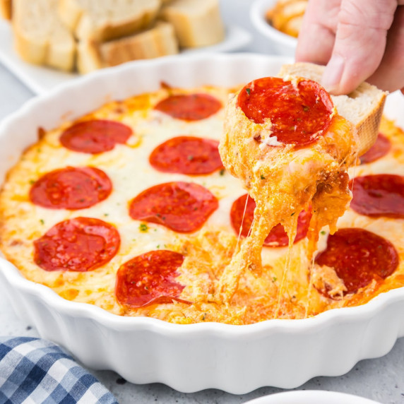A person dipping into a round pie pan of pepperoni dip with a piece of bread stretching gooey cheese