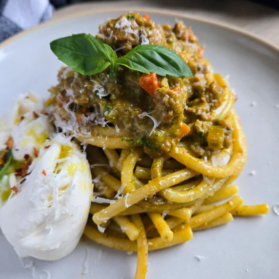 A nest of glossy bucatini topped with meat sauce and green basil alongside white burrata 