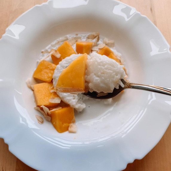 This is a white plate with fresh mango, sticky rice, coconut milk, and coconut flakes and a spoon. 