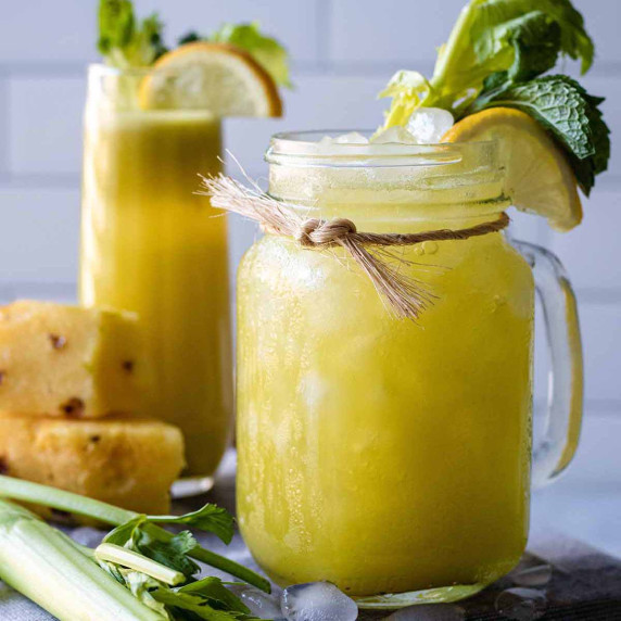 Pineapple celery juice in a glass jar mug garnished with lemon, mint leaves, and celery greens.