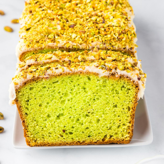 A sliced loaf of pistachio bread with green interior and white glaze, topped with chopped pistachios