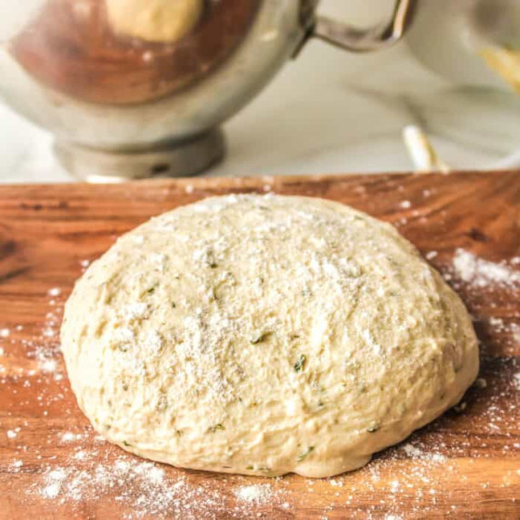 Pizza dough on a cutting board with flour and a mixer bowl behind.