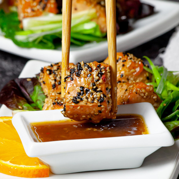 A set of chopsticks, holding a piece of orange teriyaki salmon over a sauce in a white dipping bowl.