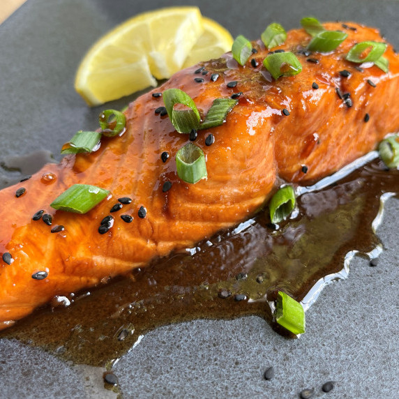 Glazed salmon filet garnished with lemon, green onion, and black sesame seeds on a gray plate.