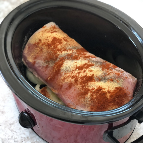 Image of a crock pot with a piece of pork loin sitting on sliced onion and covered with spices.