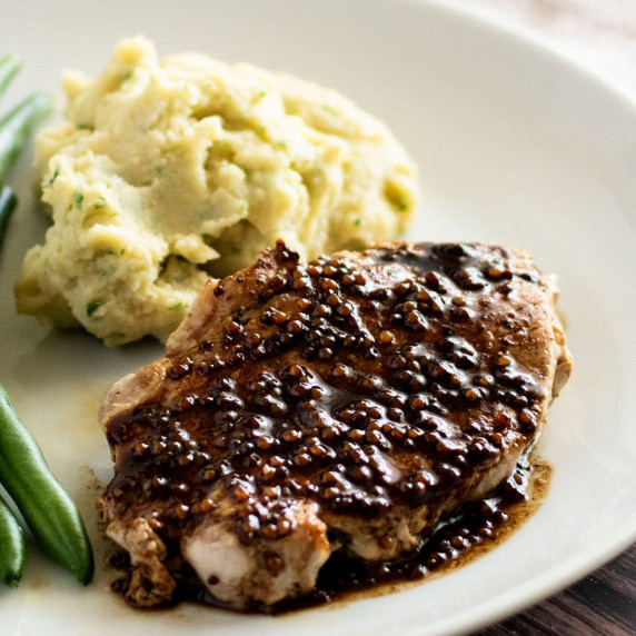 A pan fried pork steak smothered in a balsamic glaze on a dinner plate with mashed potatos & beans.