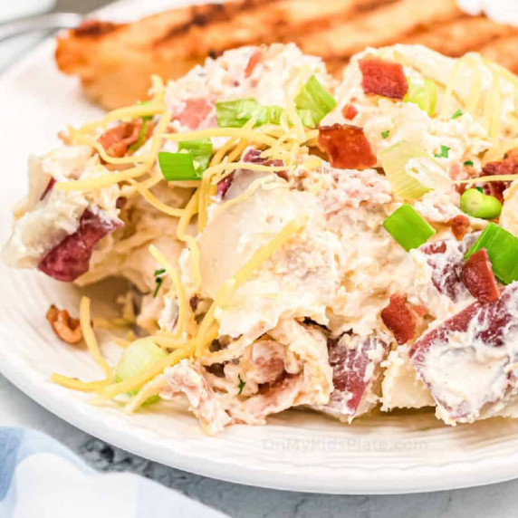 Potato salad topped with cheese, green onions and bacon on a dinner plate with chicken behind.