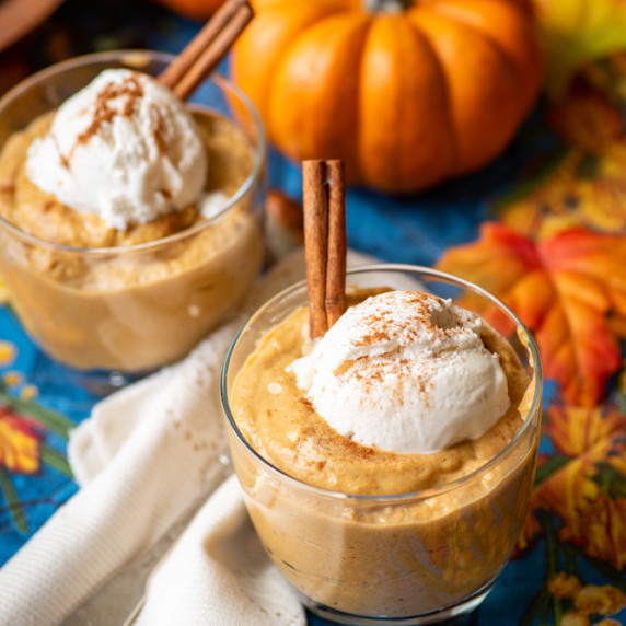 Glasses of pumpkin pudding topped with coconut whip and cinnamon sticks on a placemat.