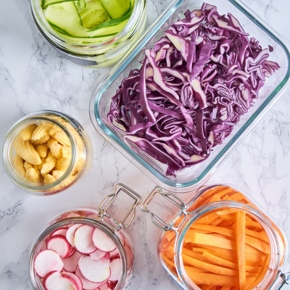 5 glass containers with pickled red cabbage, radishes, carrots, mini sweetcorn and cucumbers.