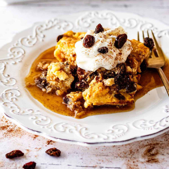 Serving of raisin bread pudding topped with whipped cream and raisins on a white plate