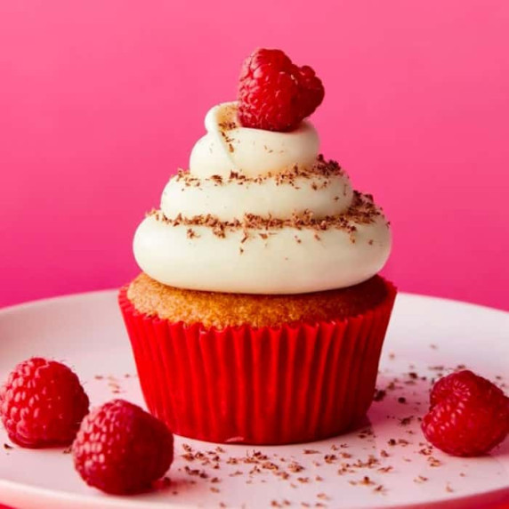 A raspberry cream cheese cupcake on a plate surrounded by scattered fresh raspberries.