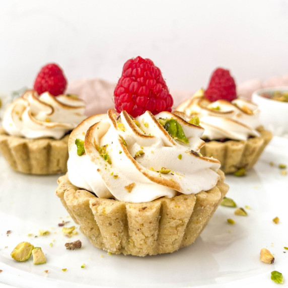 raspberry pistachio tartlet on a white plate with more tartlets in the background