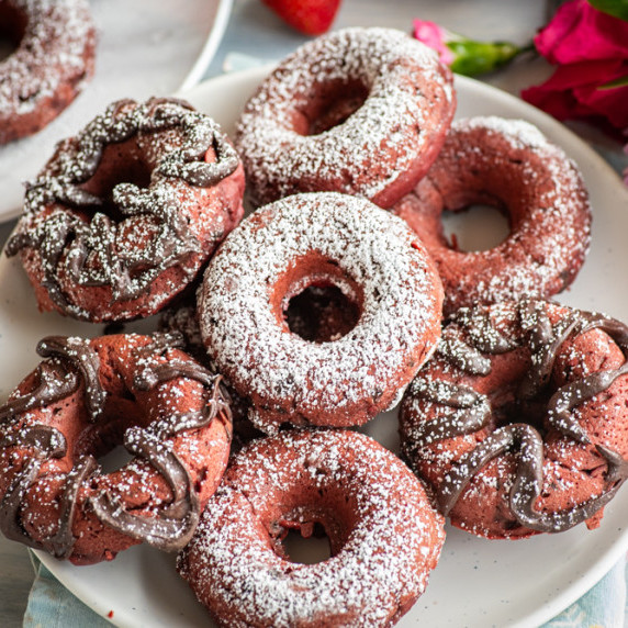 A plate of pink red velvet donuts, drizzled with chocolate or sprinkled with powdered sugar