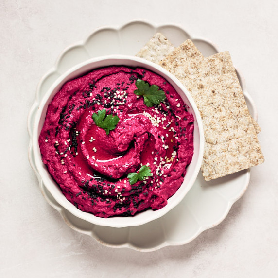 Roasted beet hummus in a white bowl on a plate with rye crackers on a grey surface