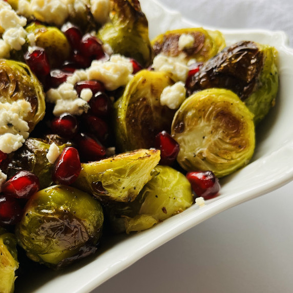 Roasted halved Brussels sprouts topped with blue cheese and pomegranate arils in a white bowl.