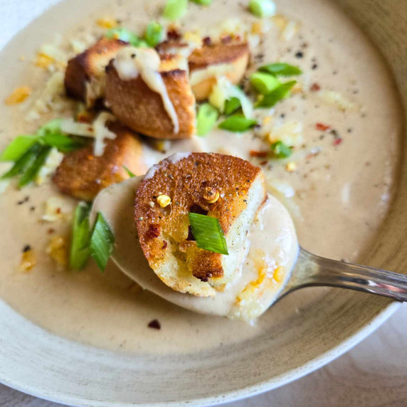 A spoon carrying creamy cauliflower soup and a golden brown, toasted crouton with pops of green