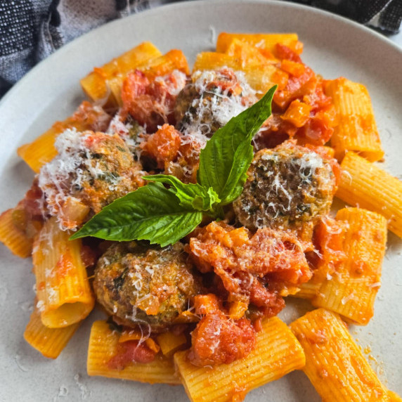 A white plate with red sauce rigatoni, meatballs, and fresh green basil.