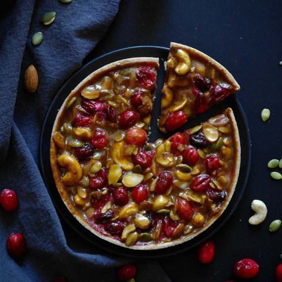 A tart filled with nuts, caramel and cranberries on a dark surface.