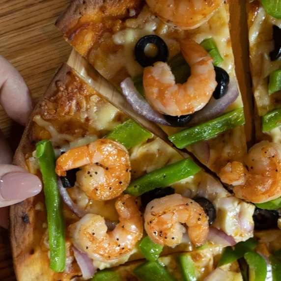 hand of a woman with pink nail polished nails grabbing a slice of shrimp pizza with green peppers
