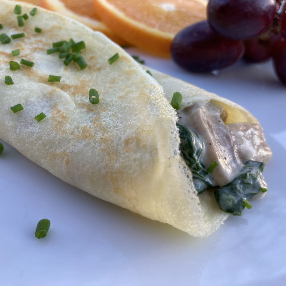 A crepe stuffed with spinach and mushrooms in a cream sauce on a white plate with fresh fruit.
